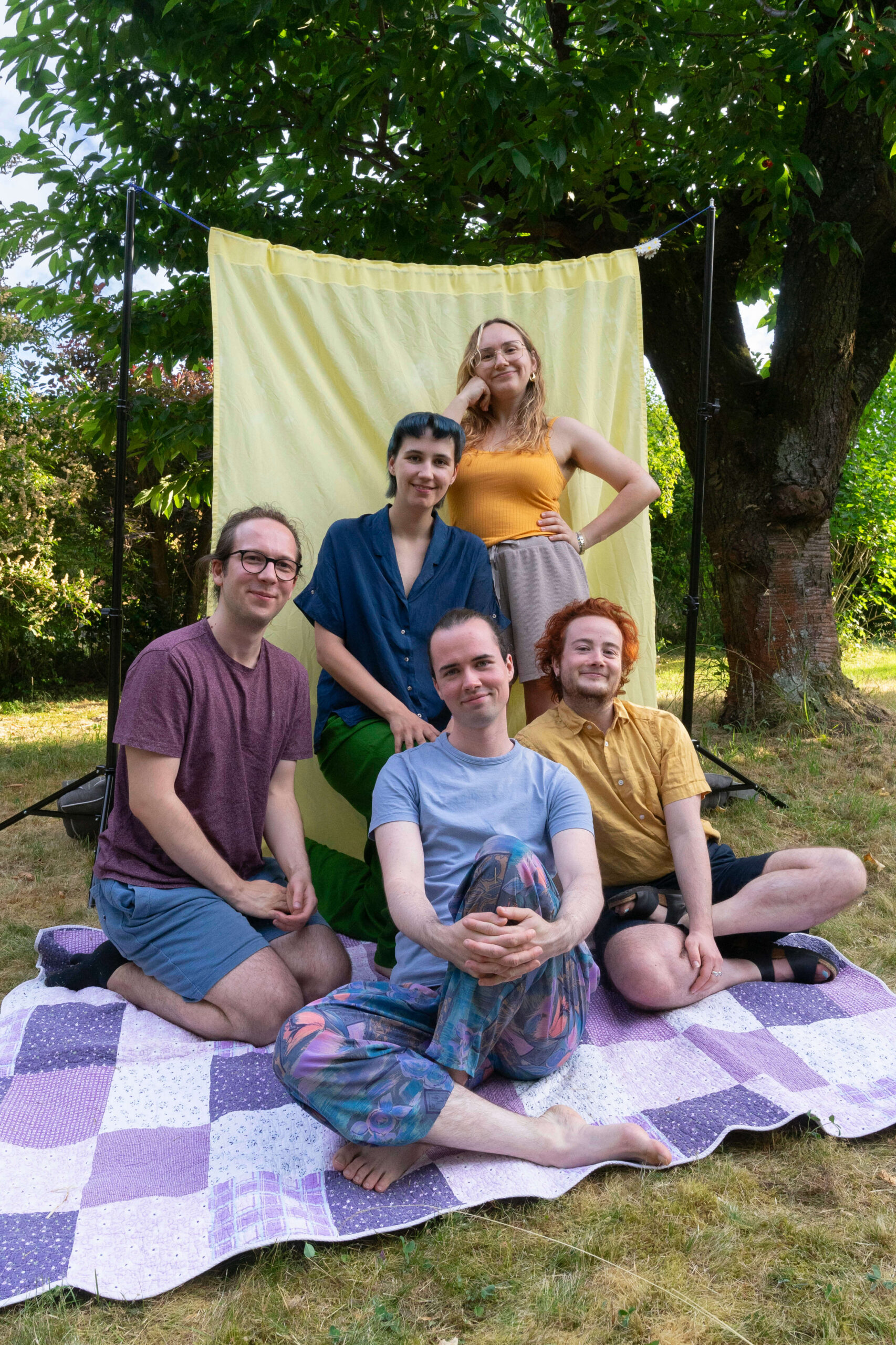 Die Queerdinx-Redaktion sitzt und steht zusammen auf einer Picknickdecke. Hinter ihnen ist ein großer Baum, der durch eine Behilfsleinwand verdeckt wird. Alle lachen in die Kamera und sind in zueinander passenden Farben (blau, hellblau, gelb und lila) gekleidet.