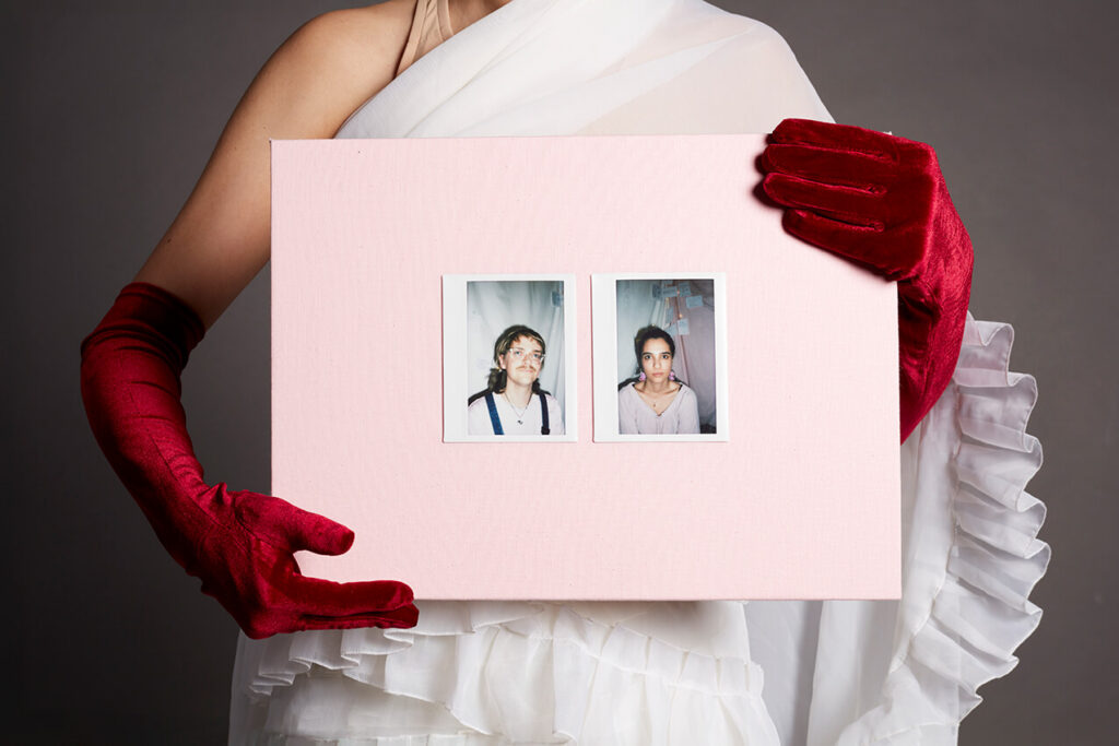Eine Person mit roten Samthandschuhen hält eine rosa Tafel. Auf dieser sind Instax-Fotografien geklebt, die weitere Personen in unterschiedlichen Posen zeigen.