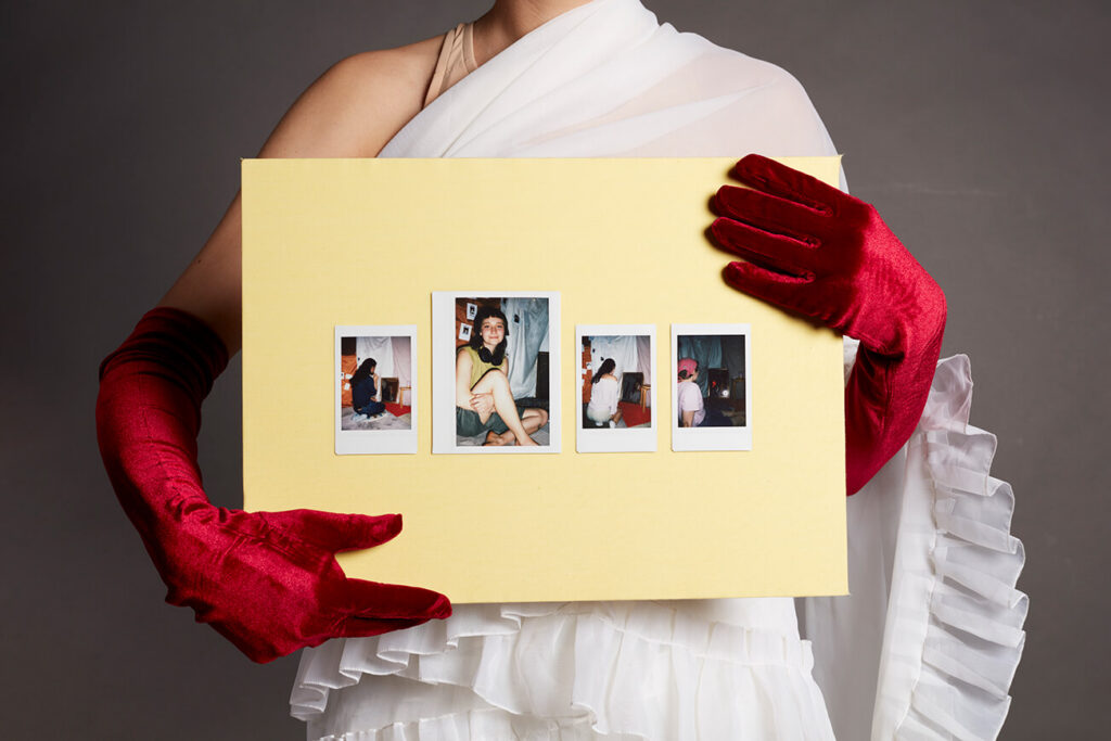 Eine Person mit roten Samthandschuhen hält eine gelbe Tafel. Auf dieser sind Instax-Fotografien geklebt, die weitere Personen in unterschiedlichen Posen zeigen.