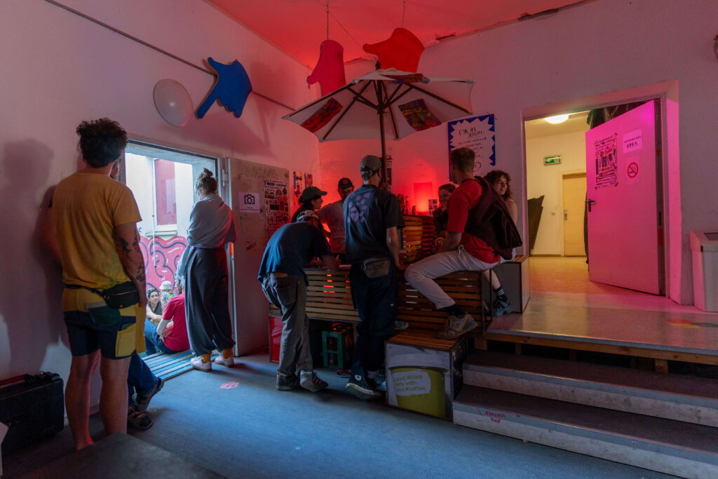 Ein Blick in einen Raum zeigt eine Bar, die rot beleuchtet ist, an der viele Menschen sitzen und stehen.