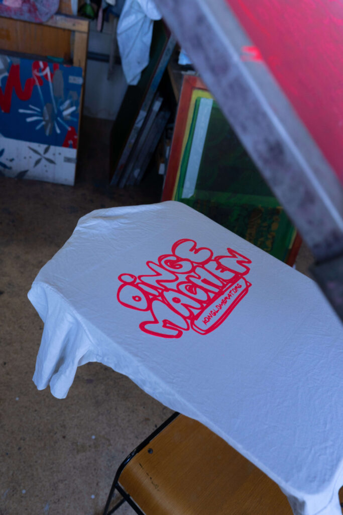 Auf einem weißen Tshirt steht in pinken Lettern "Dinge machen". Der Schriftzug wurde scheinbar soeben mit Siebdruck gedruckt, denn das Sieb mit pinker Farbe ist oben im Bildrand noch sichtbar.