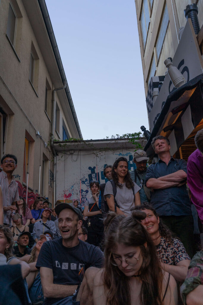 Im Innenhof: Viele Personen stehen und sitzen zusammen und schauen amüsiert in die Richtung hinter der Kamera.