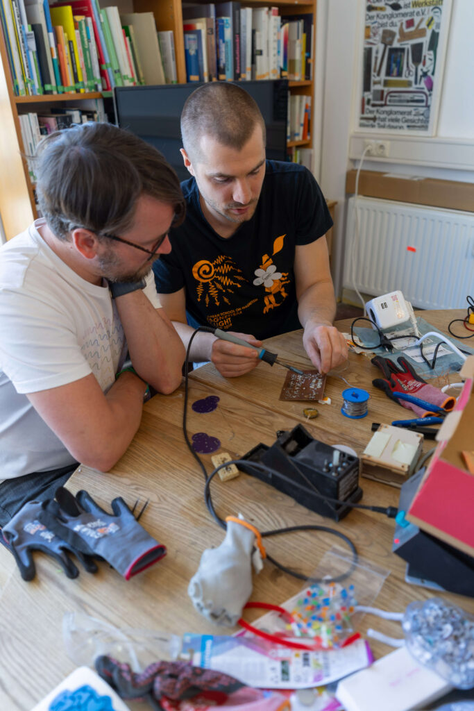 Eine Person zeigt einer anderen wie man etwas lötet. Sie sitzen an einem Tisch, auf dem viel Elektrik rumliegt.