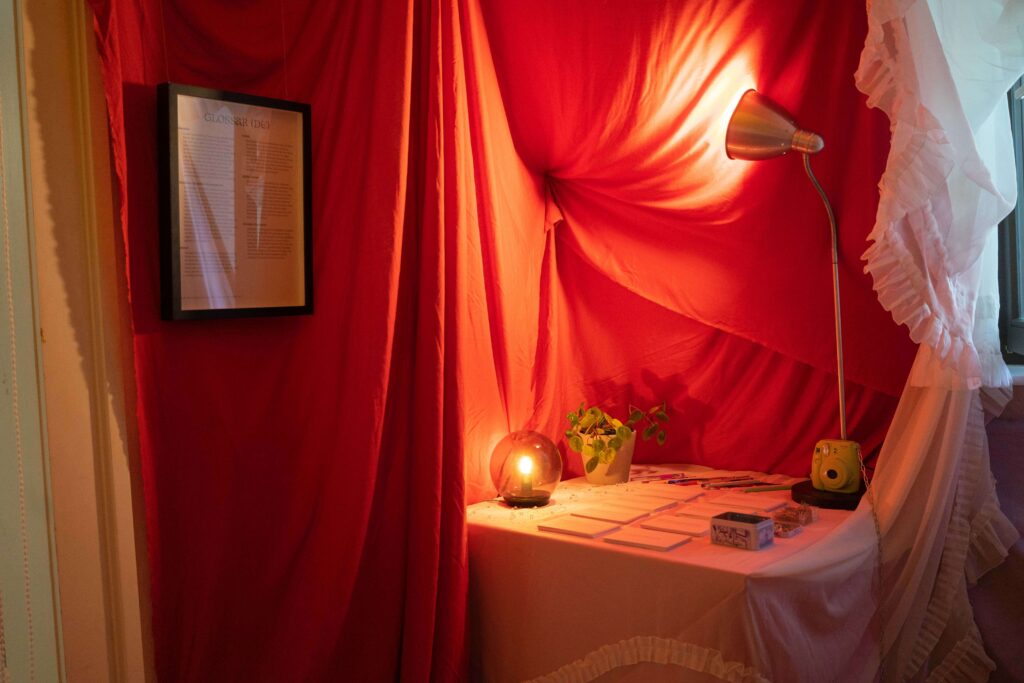 Diese Fotografie zeigt einen Tisch in einem mit Stoff behangenen Raum. Auf dem Tisch steht eine Lampe, die einen tiefroten Stoff beleuchtet. Auf dem Tisch liegen mehrere Karten sowie Stifte.