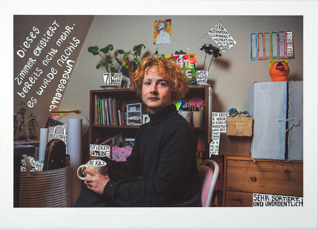 Das Bild zeigt eine Collage. Im Hintergrund ist eine Fotografie: Eine Person mit schwarzem Rollkragenpullover und rötlichen, lockigen Haaren sitzt auf einem Stuhl und schaut in die Kamera. Im Hintergrund der Fotografie sieht man das Atelier der Person mit Schränken, Tischen, usw. Als Teil der Collage wurde auf dem Foto wurde mit weißem Stift geschrieben.