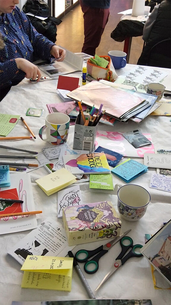 Auf einem Tisch liegen wild verteilt Zines und Bastelmaterialien wie Zeitschriften, Sticker und Scheren.